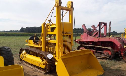 A yellow Trackson bulldozer with a front blade, parked in a field with green crops in the background.