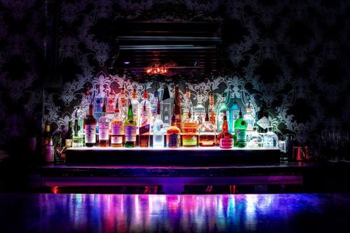 A vibrant bar shelf filled with various bottles of liquor, illuminated by colorful lights against a patterned background.