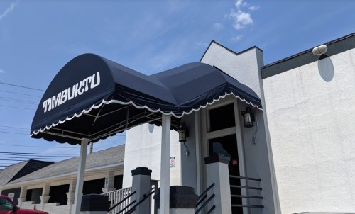 A blue awning with "TIMBUKTU" text above the entrance of a building under a clear blue sky.