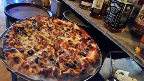 A freshly baked pizza topped with olives and meat, placed on a counter near bottles of whiskey.