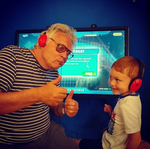 A man and a boy wearing headphones give thumbs up in front of a video game screen, smiling and enjoying their time together.