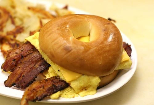 A bagel sandwich with cheese, scrambled eggs, and crispy bacon, served on a plate with hash browns in the background.