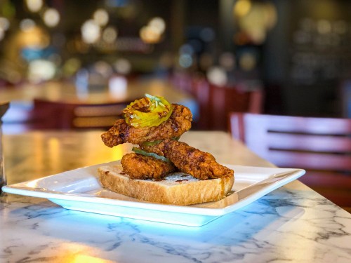 A plate with fried chicken pieces on a slice of bread, topped with pickles and green onions, set on a marble table.