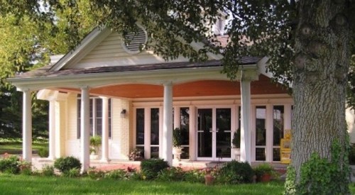A charming yellow house with a porch, surrounded by greenery and trees.