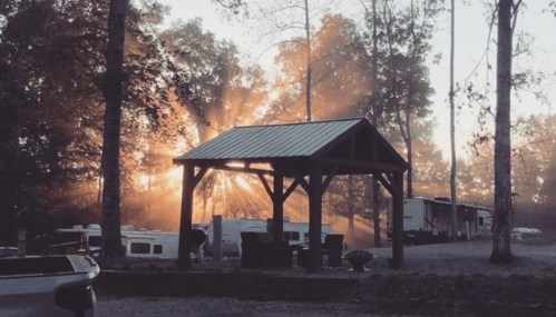 A serene campsite at dawn, with sunlight streaming through trees and illuminating a wooden pavilion.