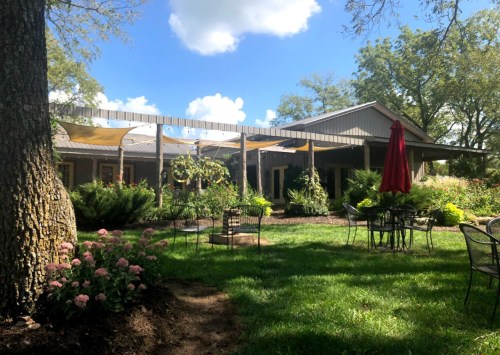 A cozy outdoor space with a garden, chairs, and a red umbrella, featuring a building with a shaded patio.
