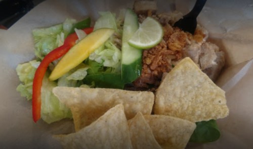 A bowl of shredded chicken topped with lettuce, mango, cucumber, lime, and crispy onions, served with tortilla chips.