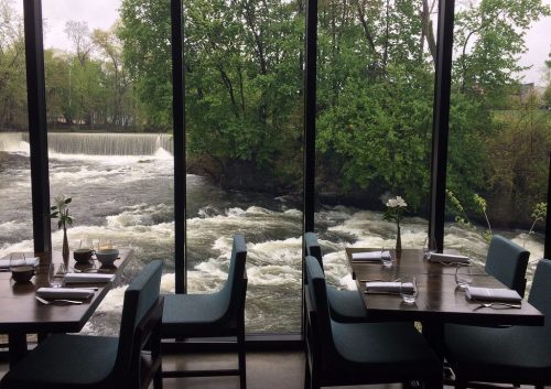 A riverside restaurant with large windows overlooking a flowing river and lush green trees. Tables are set for dining.