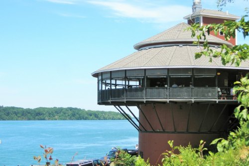 A round, modern building on stilts by a lake, surrounded by greenery and blue water under a clear sky.