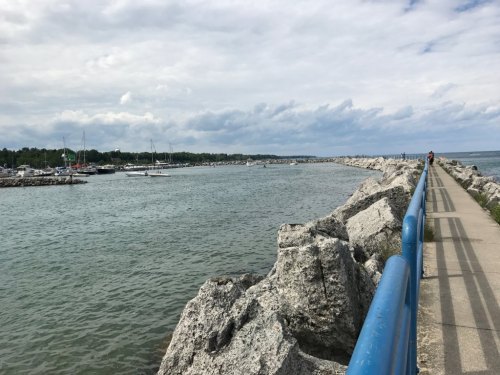 A rocky jetty extends into a calm waterway, with boats in the distance and cloudy skies overhead.