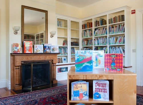 A cozy library with bookshelves, a wooden table displaying children's books, and a fireplace in the background.
