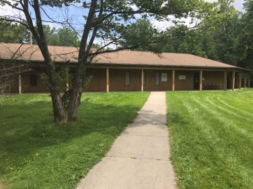 A wooden building with a sloped roof, surrounded by green grass and trees, with a concrete path leading to the entrance.