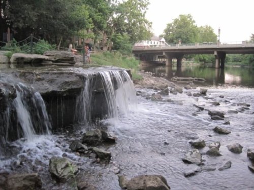 A small waterfall cascades over rocks beside a river, with trees and a bridge in the background.