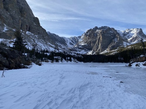 A snowy landscape with a frozen lake surrounded by mountains and evergreen trees under a clear sky.