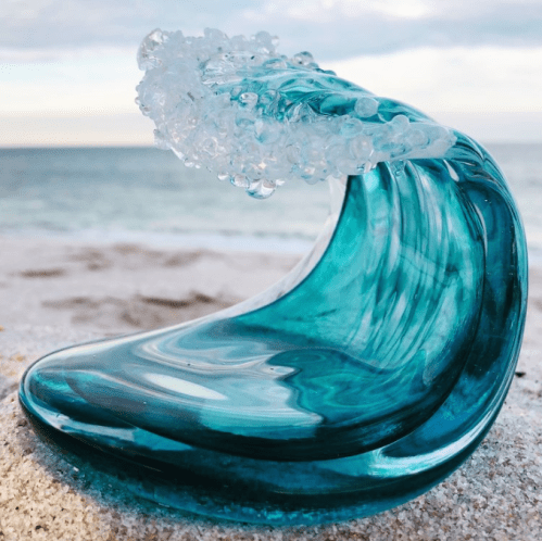 A glass sculpture of a wave, featuring turquoise and clear details, set against a beach backdrop.