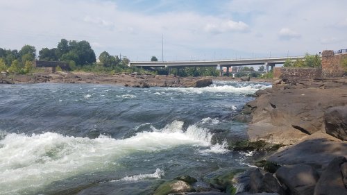 A river flows over rocky terrain under a blue sky, with a bridge visible in the background.