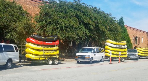 A row of stacked yellow and red rafts on a trailer next to parked vans along a brick building.