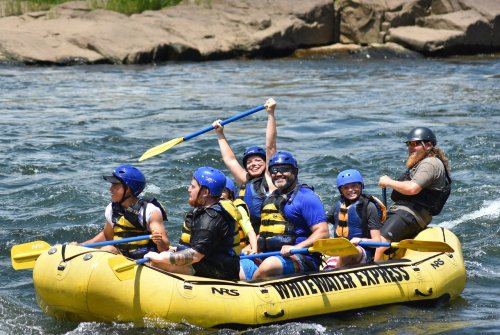 A group of six people in a yellow raft, smiling and paddling on a river, enjoying a fun white-water rafting adventure.