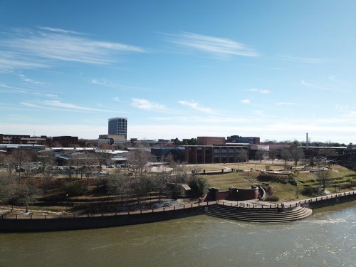 A scenic view of a riverbank park with buildings and a clear blue sky in the background.