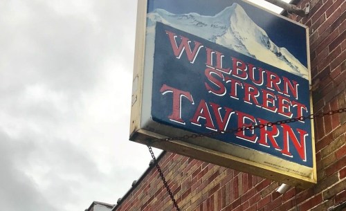 Sign for Wilburn Street Tavern with a mountain backdrop, hanging from a brick building under a cloudy sky.