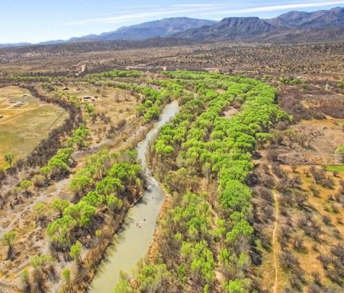 Aerial view of a winding river surrounded by lush green trees and arid landscape, with mountains in the background.
