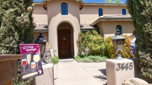 A charming adobe-style house with a sign for "Group Therapy" featuring wine glasses, surrounded by greenery.