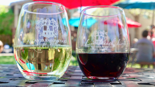 Two wine glasses on a table: one filled with white wine and the other with red, set against a colorful outdoor backdrop.