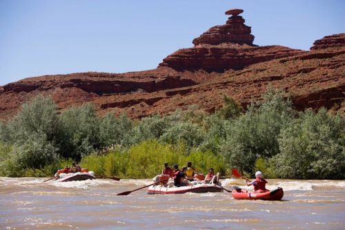 A group of people rafting on a river with red rock formations and greenery in the background under a clear blue sky.