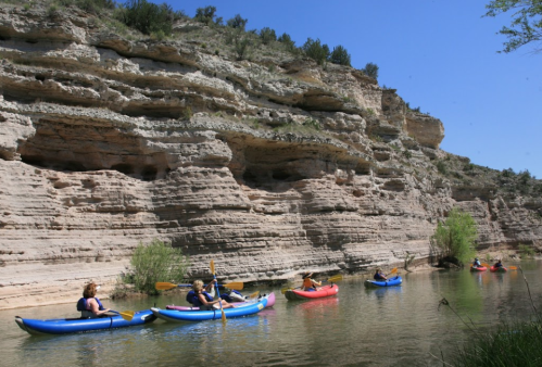 Kayakers paddle along a river with steep, rocky cliffs and greenery under a clear blue sky.