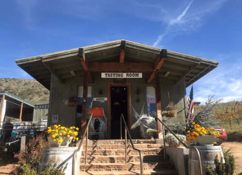 A rustic tasting room with a welcoming entrance, surrounded by flowers and mountains under a blue sky.