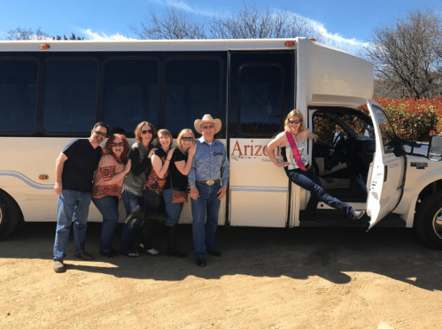 A group of six people poses playfully by a bus labeled "Arizona," with one person in a cowboy hat and sash.