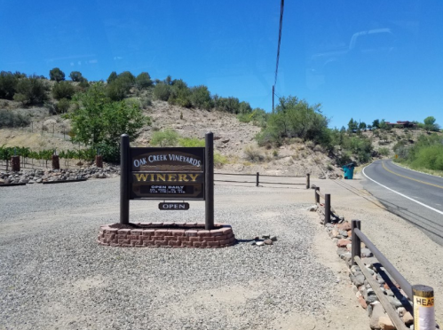 Sign for Oak Creek Vineyards Winery, indicating it's open daily, with a scenic road and greenery in the background.