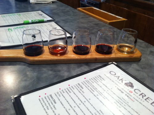 A wooden flight board with five small glasses of different colored liquids, placed on a table with menus.