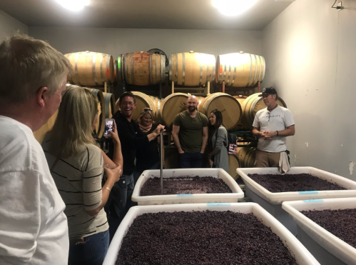 A group of people smiles and interacts in a winery, surrounded by barrels and large containers of grapes.