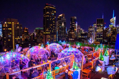 Rooftop scene at night with illuminated igloos, festive lights, and a skyline view of tall buildings.