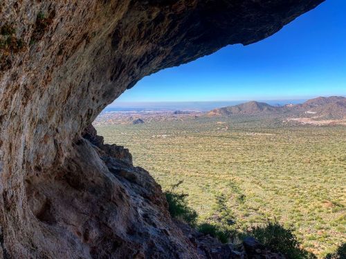 A scenic view from a rocky overhang, overlooking a vast green landscape and distant mountains under a clear blue sky.