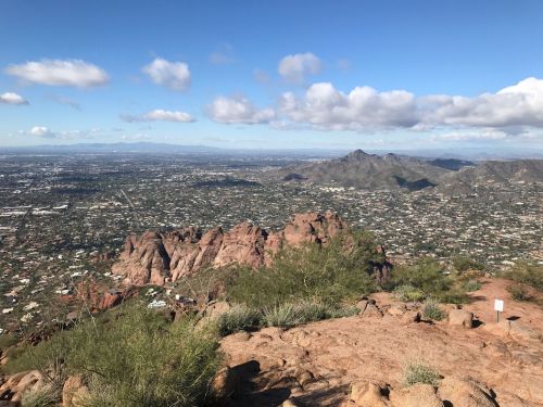 A panoramic view from a rocky mountain peak overlooking a valley with scattered clouds and distant mountains.