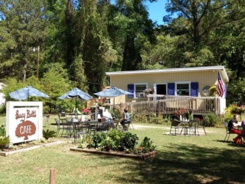 A charming café with outdoor seating, surrounded by greenery and featuring a sign that reads "Busy Bubbles Café."