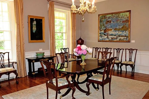 Elegant dining room with a dark wooden table, floral centerpiece, and antique chairs, featuring artwork and a chandelier.