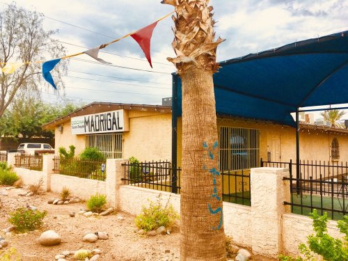 A building with a sign reading "Madrigal," surrounded by a garden and colorful flags, under a cloudy sky.