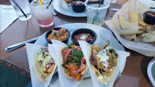 A plate of three tacos with various toppings, accompanied by rice, beans, and tortilla chips. Drinks in the background.