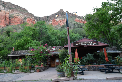 A rustic store with a sign for groceries and cold drinks, surrounded by greenery and red rock mountains in the background.