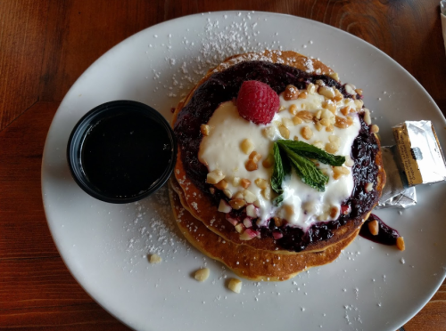Stack of pancakes topped with berry compote, whipped cream, nuts, a raspberry, and mint, served with syrup on the side.