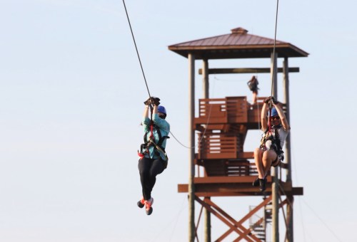 Two people ziplining in front of a wooden observation tower on a clear day.