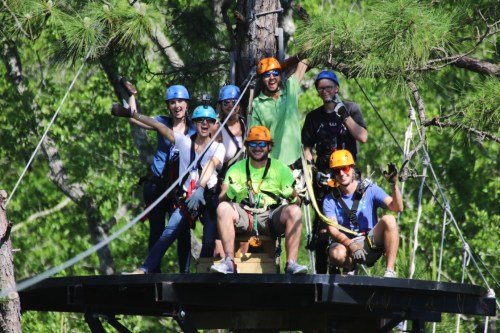 A group of eight people in helmets and harnesses, smiling and posing on a platform in a forested area.
