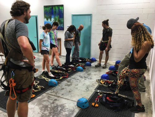 A group of people in climbing gear prepares for an activity, surrounded by helmets and equipment in a training space.