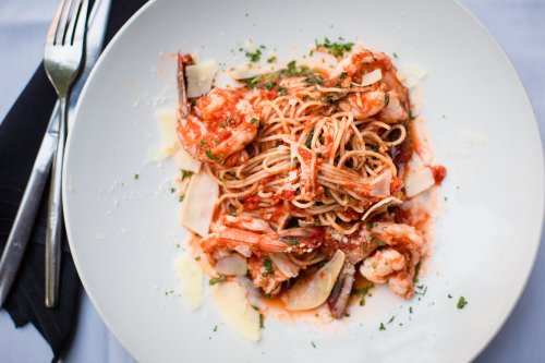 A plate of spaghetti with shrimp, garnished with parsley and cheese, served on a white dish with silverware beside it.