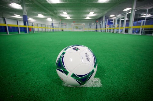 A soccer ball on a green turf field, with goalposts visible in the background of an indoor facility.