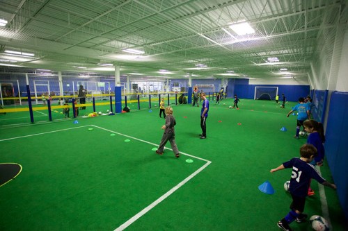 Children practice soccer indoors on a green turf field, with cones and goals set up for training activities.