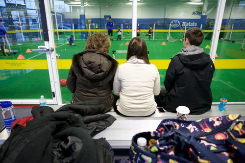 Three people sit on bleachers watching a youth soccer practice through a glass wall in a sports facility.